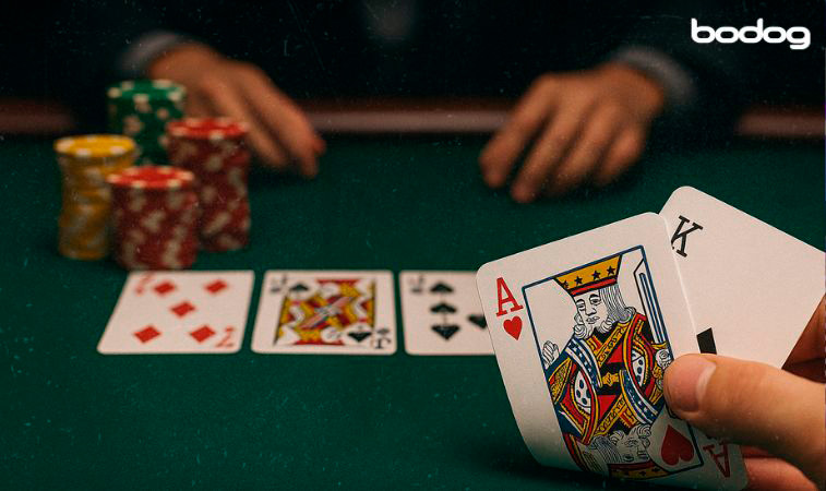  mano con cartas A-K en primer plano, cartas de un turn, fichas y manos de otra persona en segundo.